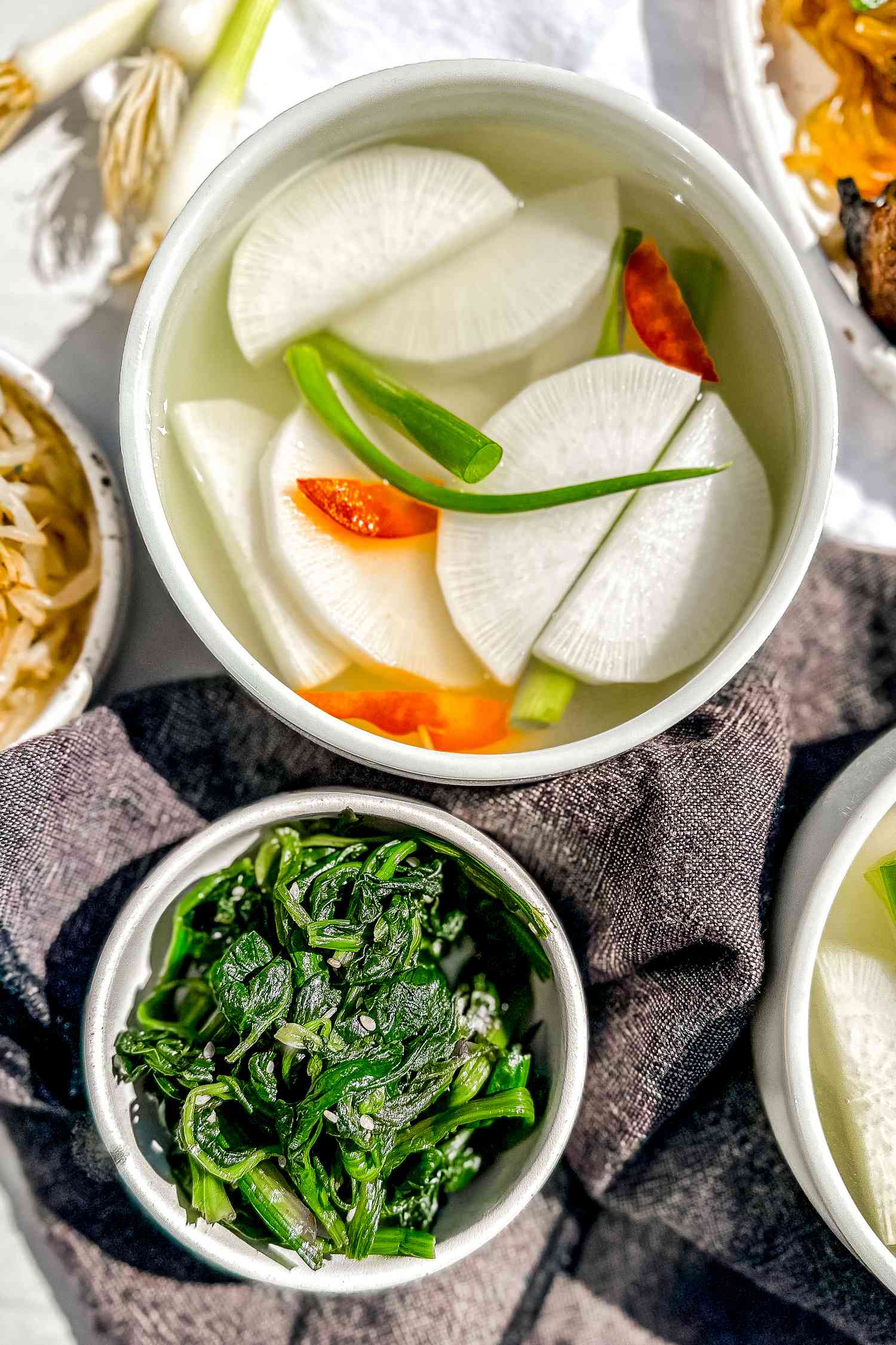 Bowl of Dongchimi Surrounded by Bowls of Banchan (Side Dishes) and a Bowl of Korean Short Ribs. On the Table, a Table Napkin and Some Whole Green Onions.