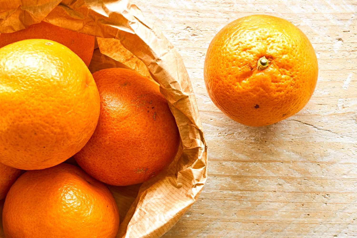 Overhead shot of a brown paper bag with oranges, and one orange sitting next to the bag on a wooden counter