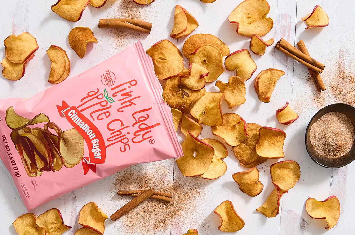 Overhead view of a bag of Trader Joe's Cinnamon Sugar Pink Lady Apple Chips with loose chips spread out on a white tabletop along with cinnamon sticks and a small bowl of cinnamon sugar