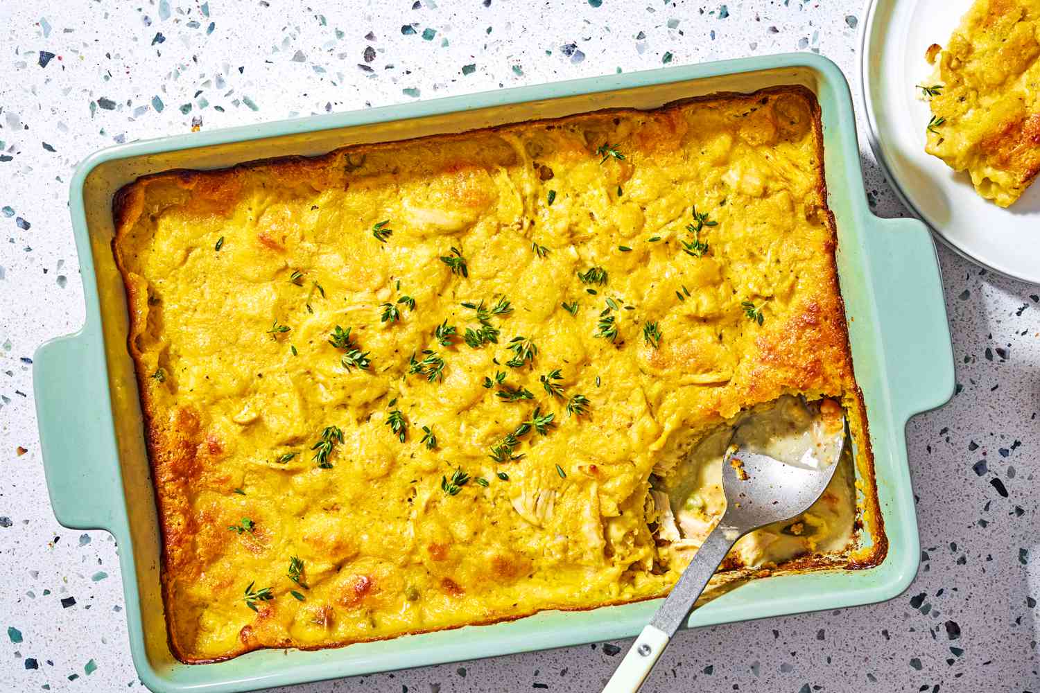 Overhead view of a green baking dish of chicken cobbler and serving spoon on a speckled countertop