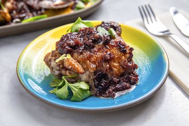 A plated chicken dish topped with a sticky sauce and garnished with herbs with a fork and knife visible in the background