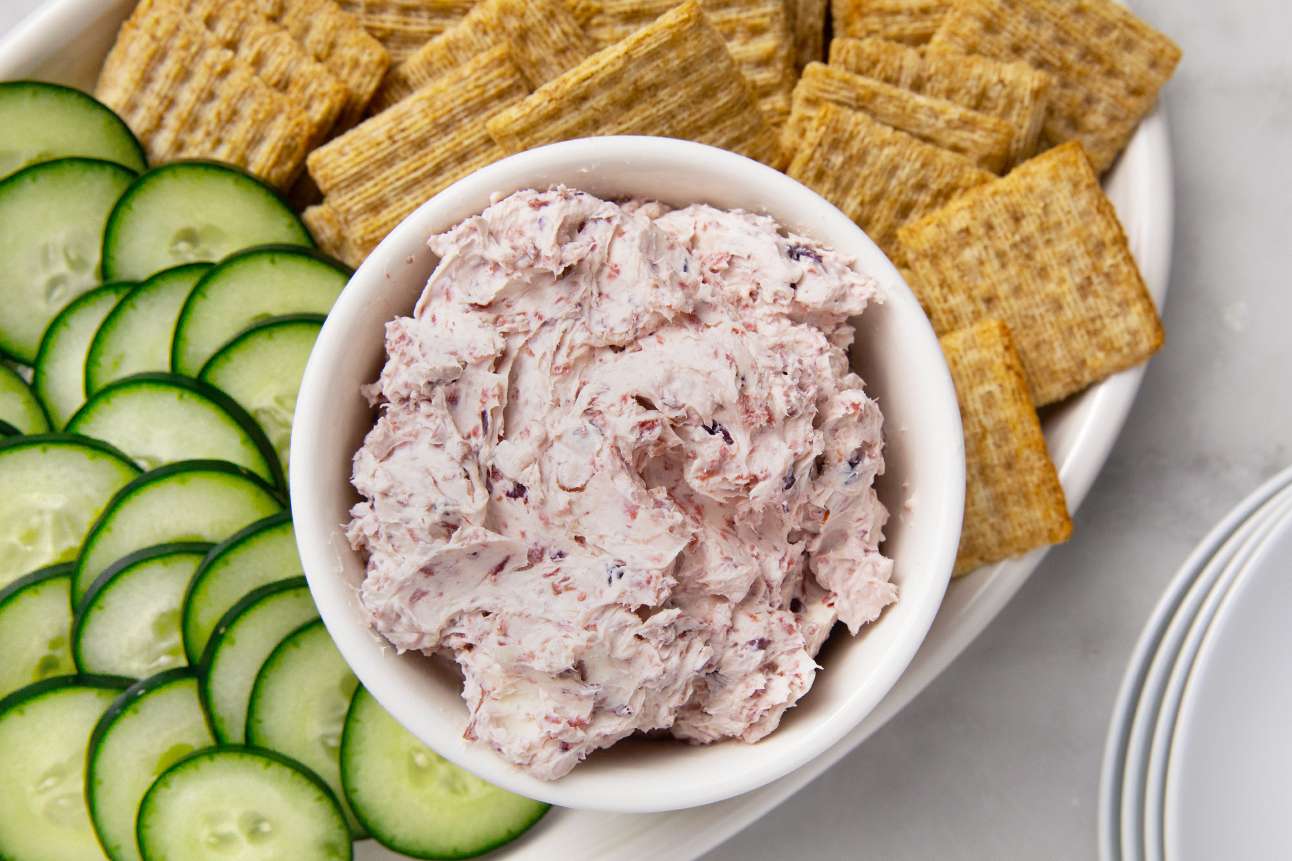 A bowl of cream cheese spread surrounded by crackers and sliced cucumbers on a platter