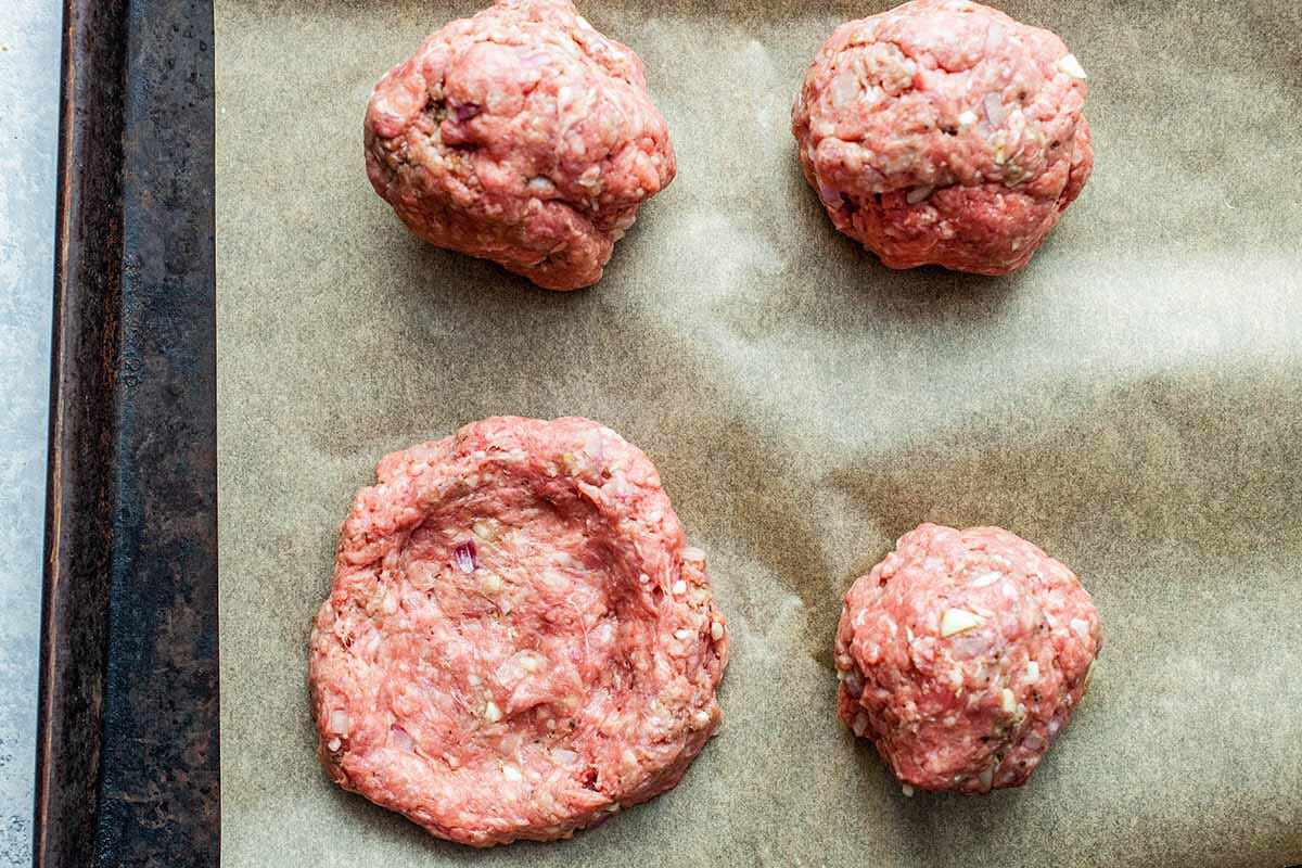 Three ground beef balls on a sheet of parchment paper and one ball that has been smashed down into a patty