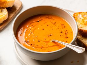 A bowl of tomato and red lentil soup with a spoon served with bread slices on the side