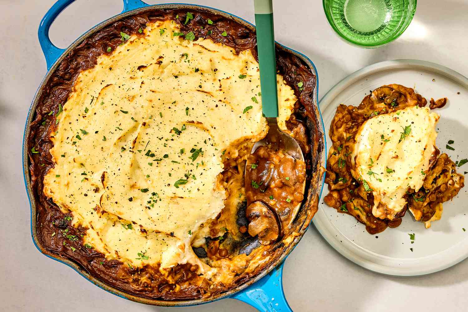 Overhead shot of lentil shepherd's pie in a skillet, with a serving spoon having scooped a serving of the dish out on a plate to the right of the skillet