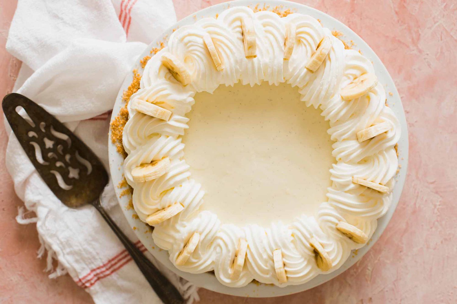Whole banana cream pie with whipped cream swirls on top and banana slices set against a pink background.