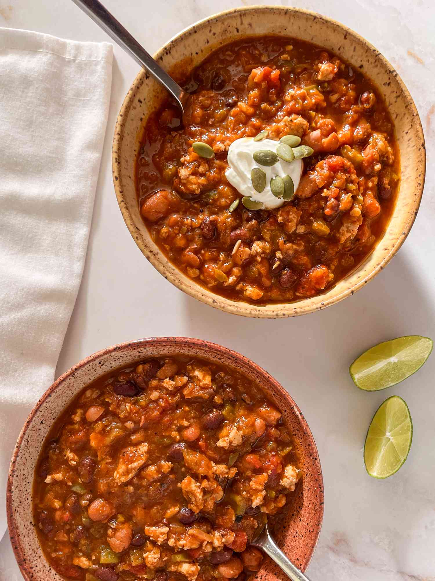 Bowl of easy pumpkin chili topped with a dollop of sour cream and pumpkin seeds at a table setting with another bowl of chili, lime wedges on the counter, and white table napkins