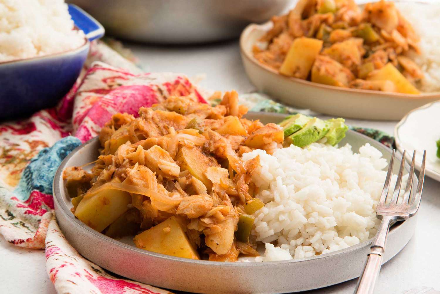 Horizontal view of rimmed grey plate with heaping portion of Puerto Rican Cod Fish. White rice and sliced avocado are on the plate along with a fork. Another plate is to the upper right and a dutch oven is partially in view in the upper center. A blue casserole dish is heaped with white rice and is above the plate to the left. A pink and blue patterened linen is under the plate and rice dish.