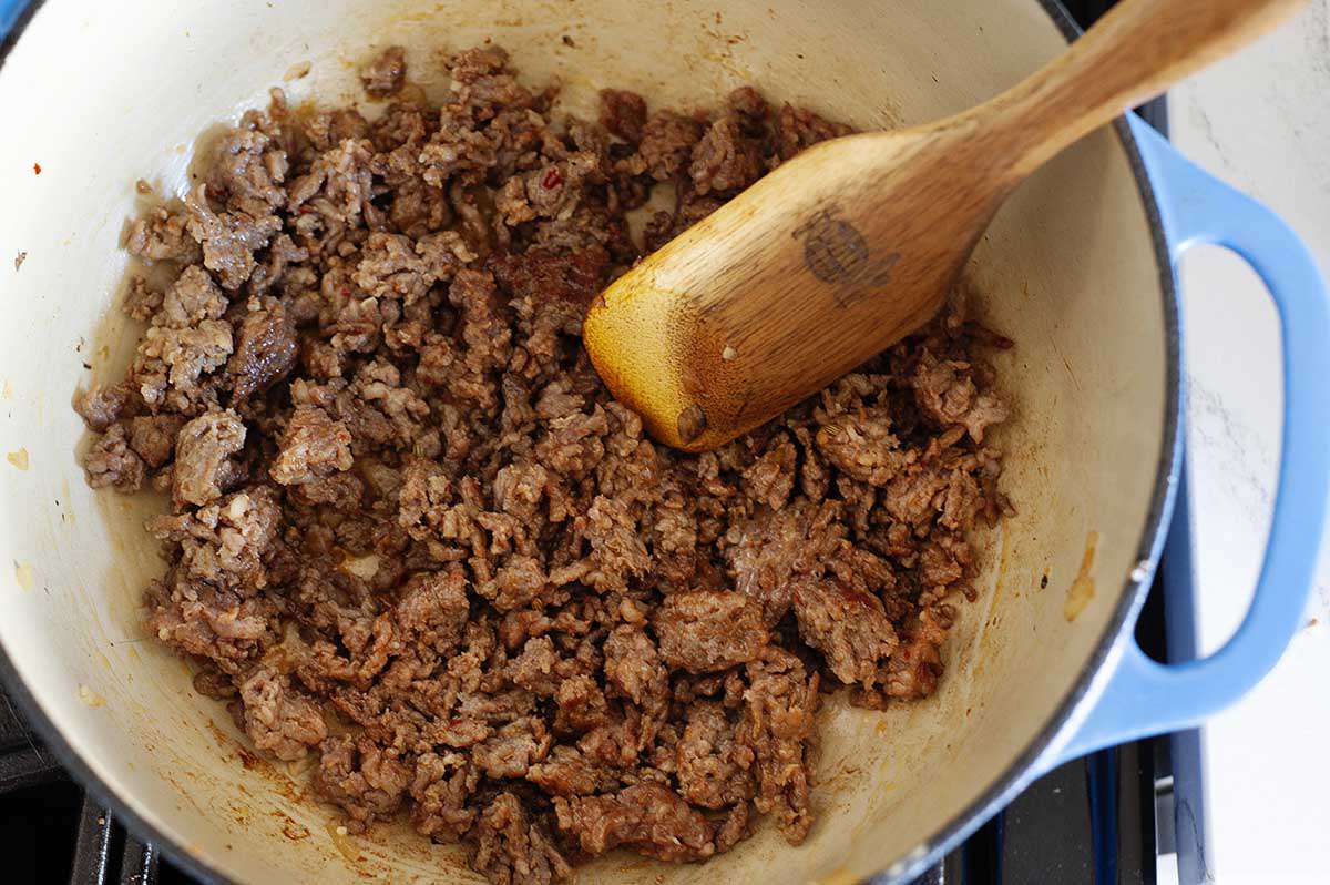 Dutch oven with crumbled sausage browning and a wooden spoon stirring it.