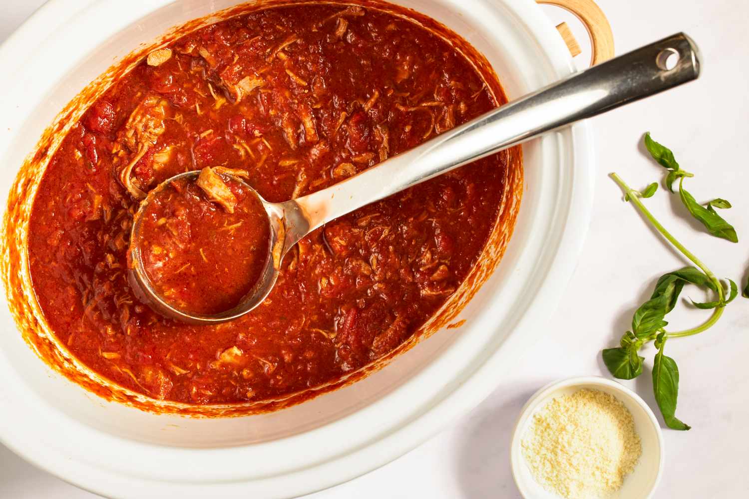 Large pot of red tomato sauce with a serving ladle garnished with fresh herbs and a small bowl of grated cheese beside it