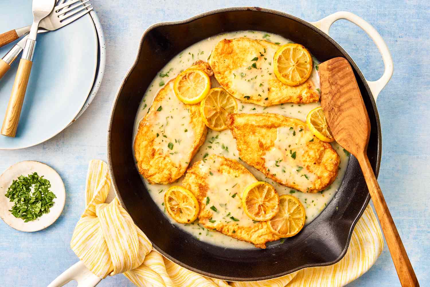 chicken francese recipe in a cast iron skillet with a wooden spatula on a table with a stack of plates with forks and a small bowl of minced herbs