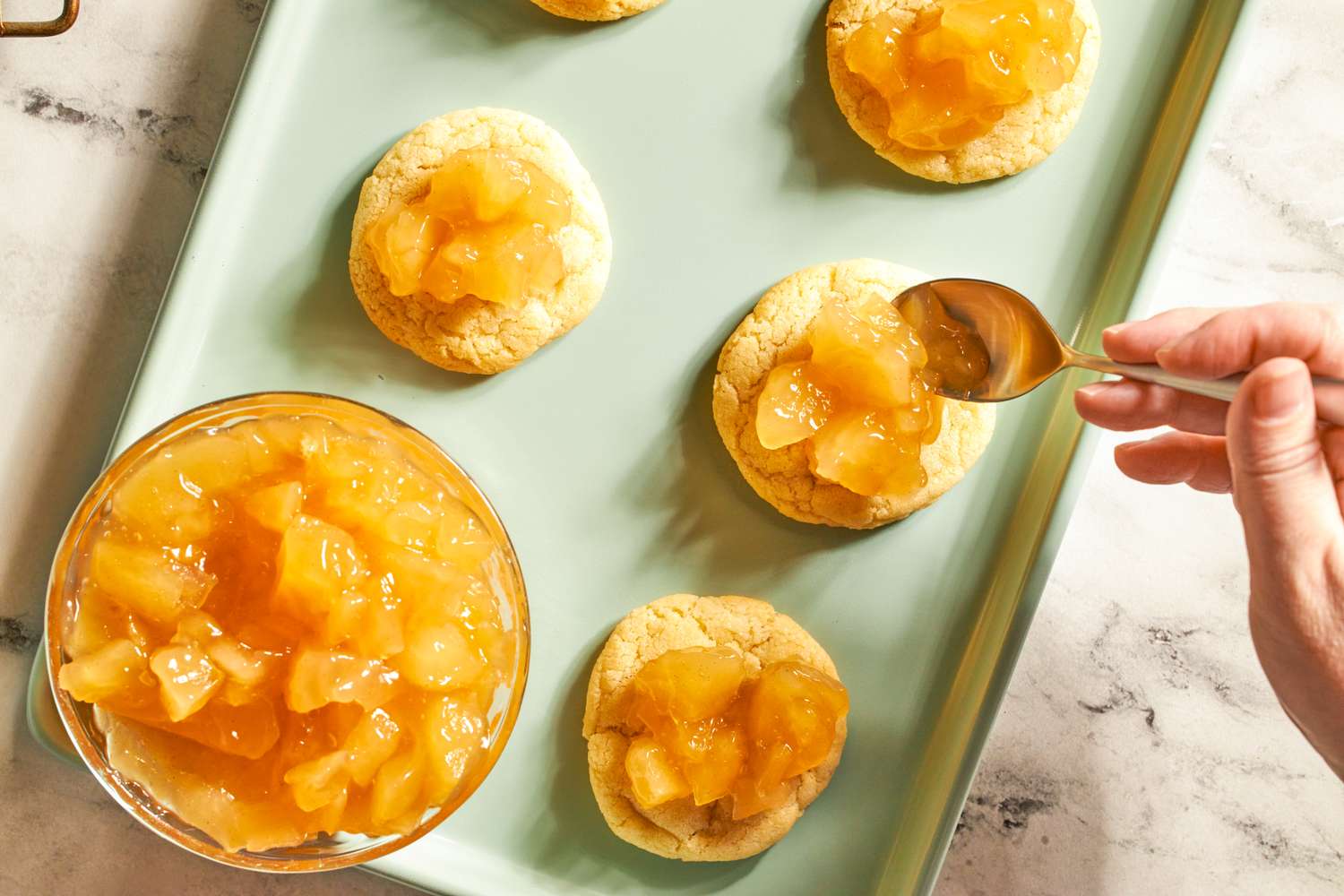 A tray of cookies being topped with apple filling using a spoon