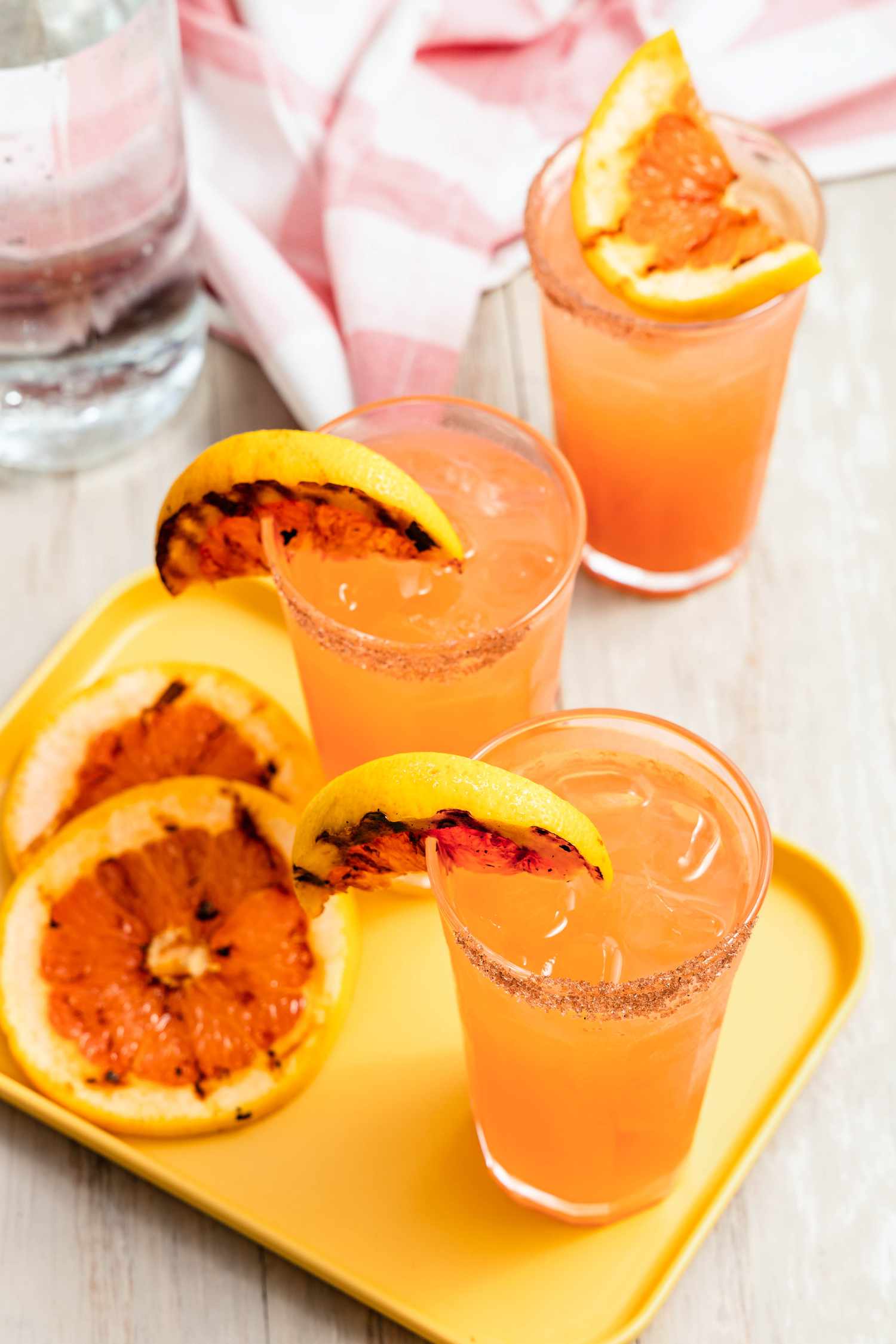 Two Smoked Salt Rimmed Glasses of Grilled Grapefruit Paloma With Two Slices of Grilled Grapefruit on the Glass Rim Next to More Slices of Grilled Grapefruit on a Small Tray, and in the Surroundings, a Bottle of Club Soda, Another Glass of Paloma, and a Pink and White Kitchen Towel