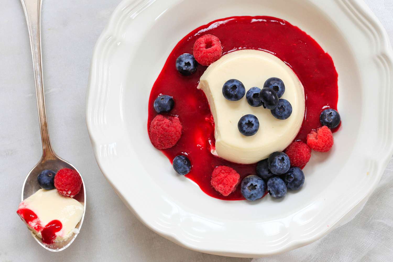 Panna Cotta Topped With Summer Berries and a Berry Sauce in a Bowl, and on the Counter a Spoonful of Panna Cotta With a Raspberry and Blueberry