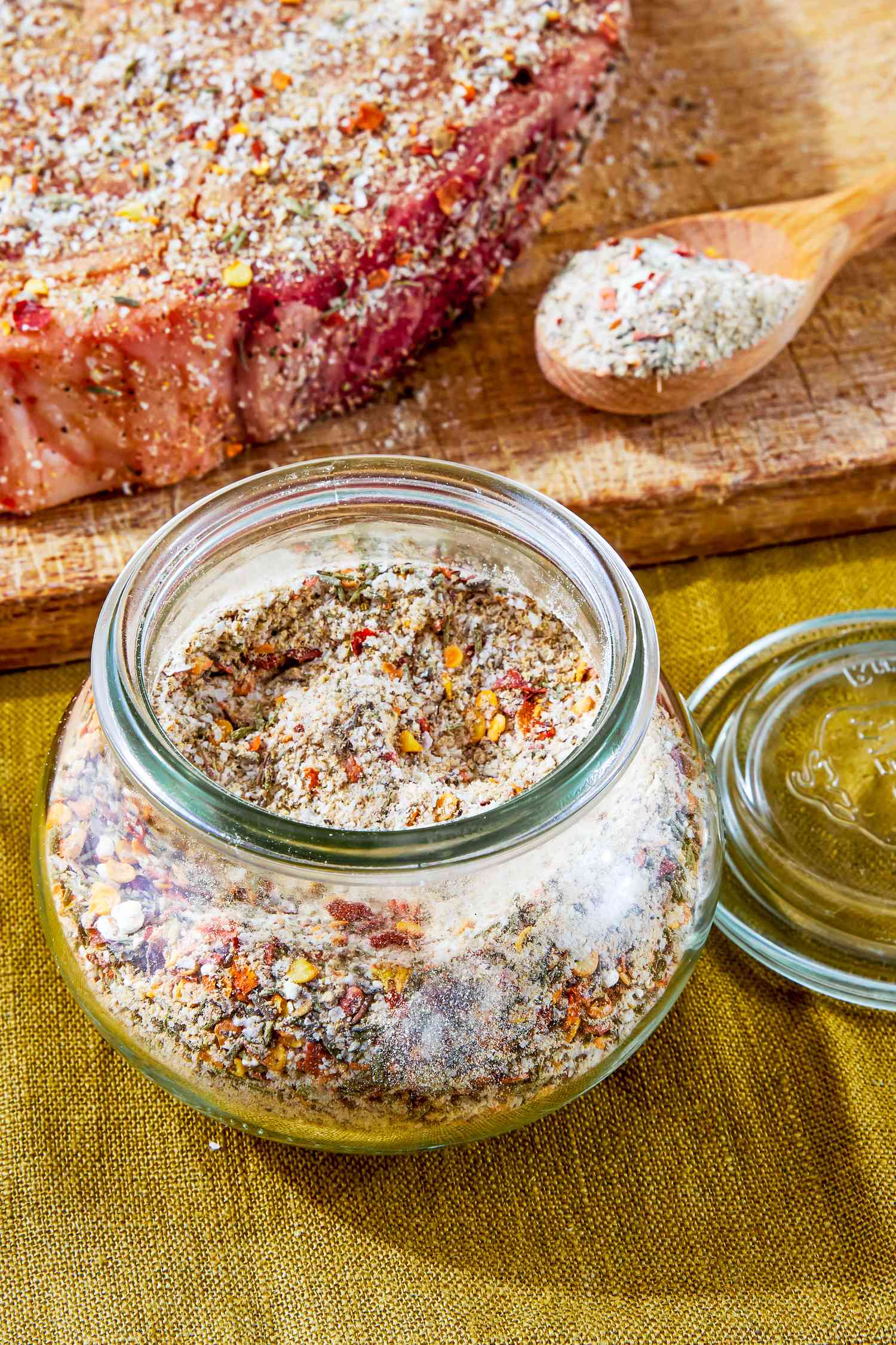 Jar of Steak Seasoning on the Counter, and in the Background, a Raw Seasoned Steak and a Spoon With More Seasoning on a Cutting Board