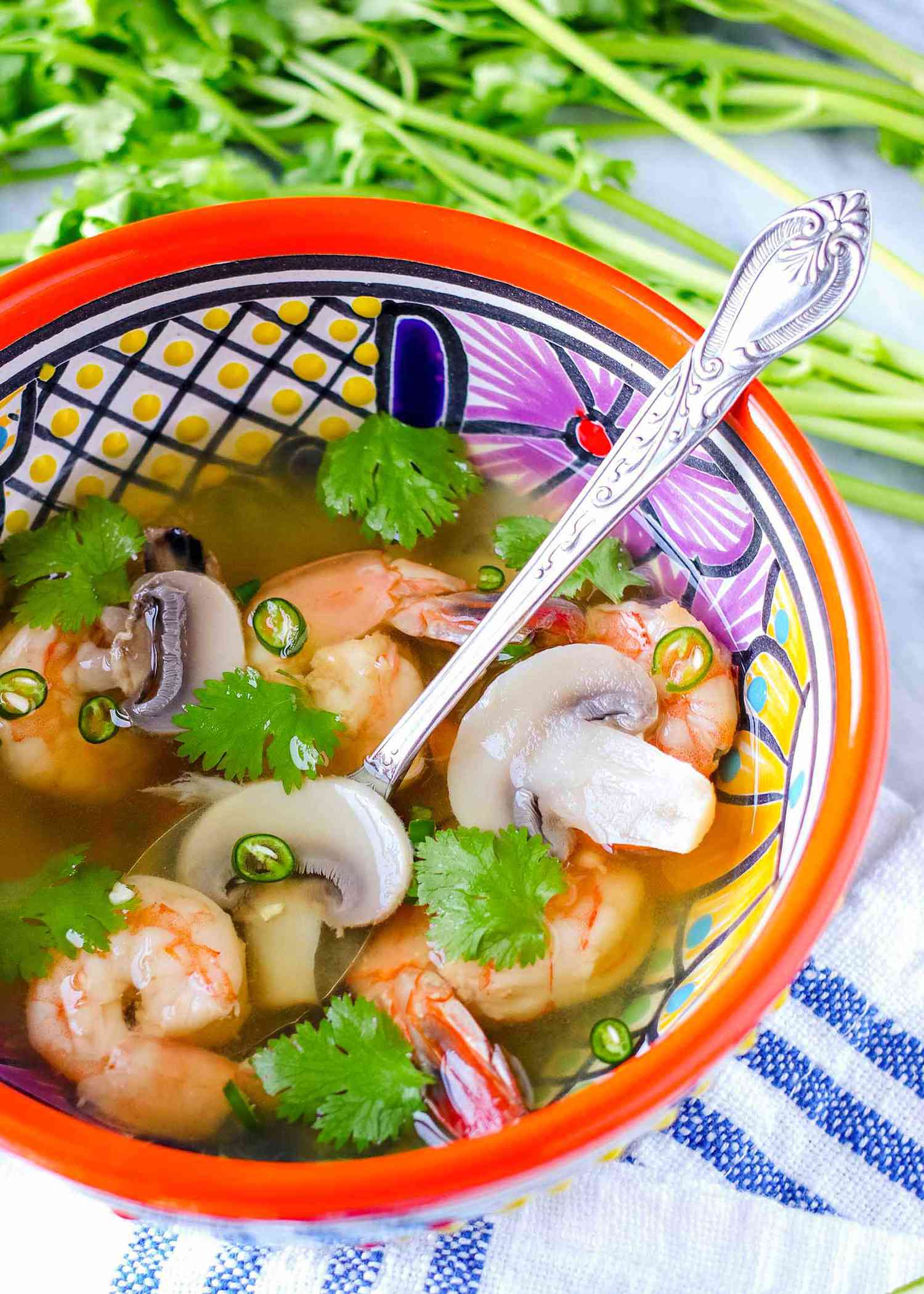 Vertical view of brightly patterned bowl with a silver spoon inside. Tom yum soup with shrimp, cilantro and mushrooms in a light broth are in the bowl. Above the bowl are stems of cilantro and below the bowl is a blue striped linen.