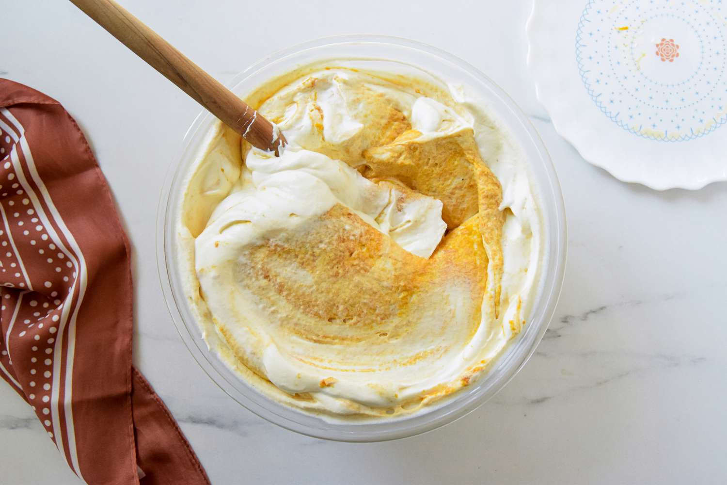 Whipped cream and pumpkin puree continiously folded together using a spatula in a bowl next to a browl kitchen linen