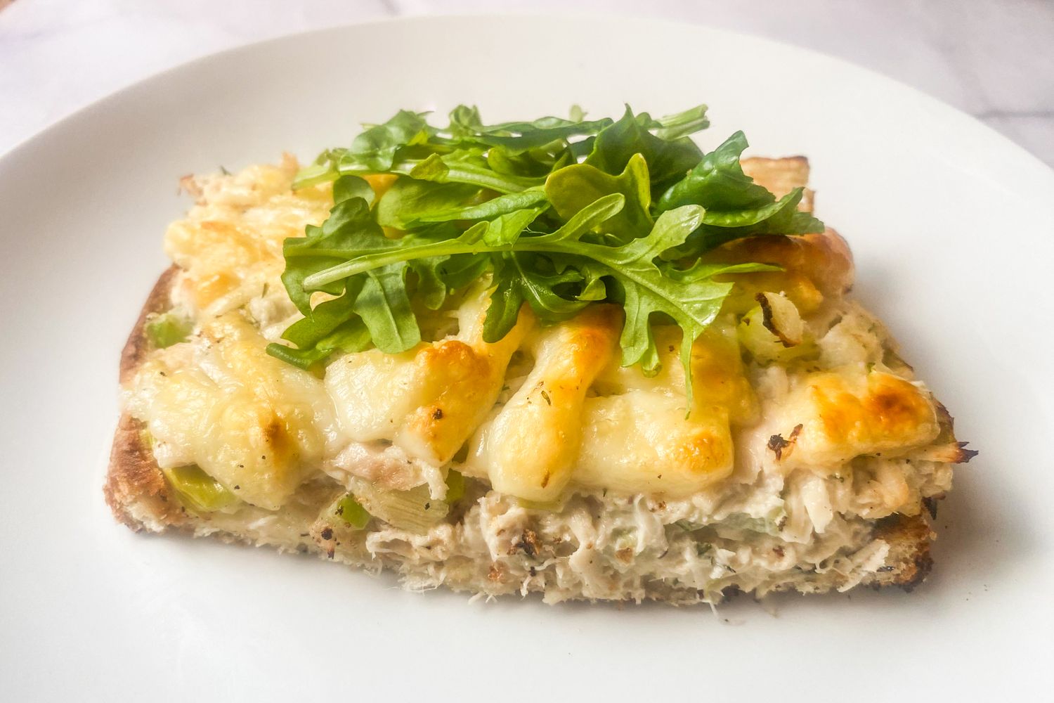 An open-faced tuna melt topped with melted cheese and arugula on a white plate