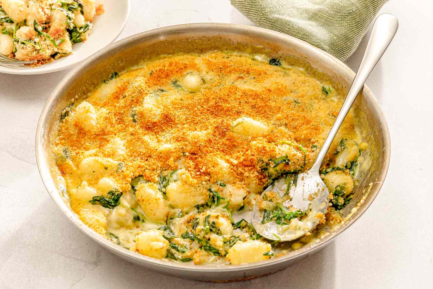 A dish of baked gnocchi with spinach and a golden breadcrumb topping served in a pan with a portion scooped into a small bowl