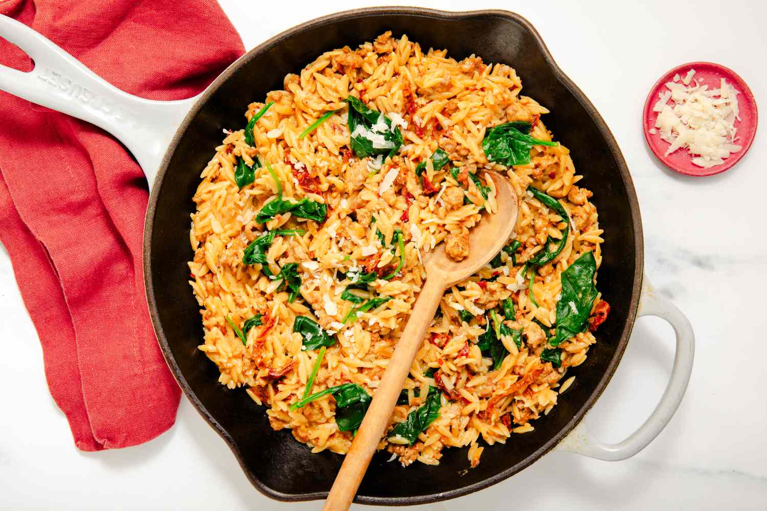 Overhead view of a skillet of orzo, italian sausage, sun-dried tomatoes and baby spinach with a wooden serving spoon