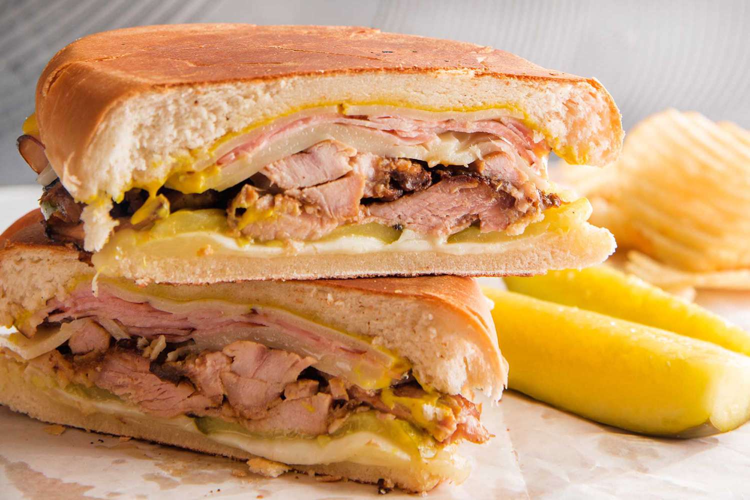 A grilled cuban sandwich is stacked on top of each other with shredded pork, mustard, cheese and pickles visible. Pickle spears and ridged potato chips are to the right of the sandwich.