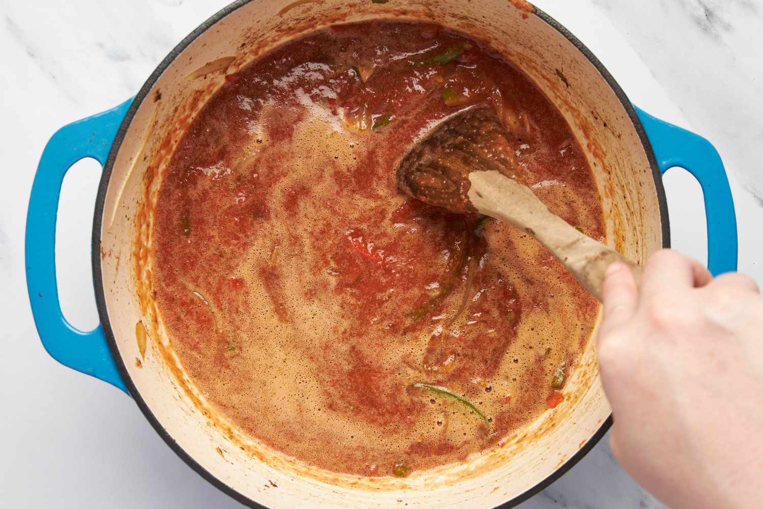 Sauce stirred with the cooked onions and bell peppers in the Dutch oven using a wooden spoon for Thinly sliced onions and bell peppers cooked in a Dutch oven for carne picada recipe