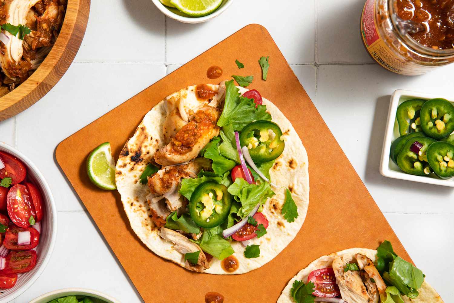 Tacos with chicken, lettuce, and jalapeños on a cutting board, surrounded by garnishes and toppings