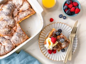 Overhead view of a white baking dish of Croissant French Toast casserole with a slice removed and placed on a small plate with a knife and fork along with a glass of orange juice and bowl of fruit