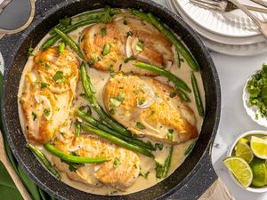 creamy coconut lime chicken in a pan at a table setting with a stack of plates, a bowl of minced cilantro, and a bowl of lime wedges