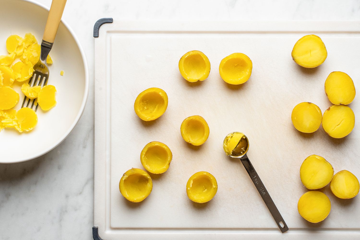 Halved and hollowed potatoes with removed insides on a cutting board with a scooping spoon and a bowl holding the scooped contents