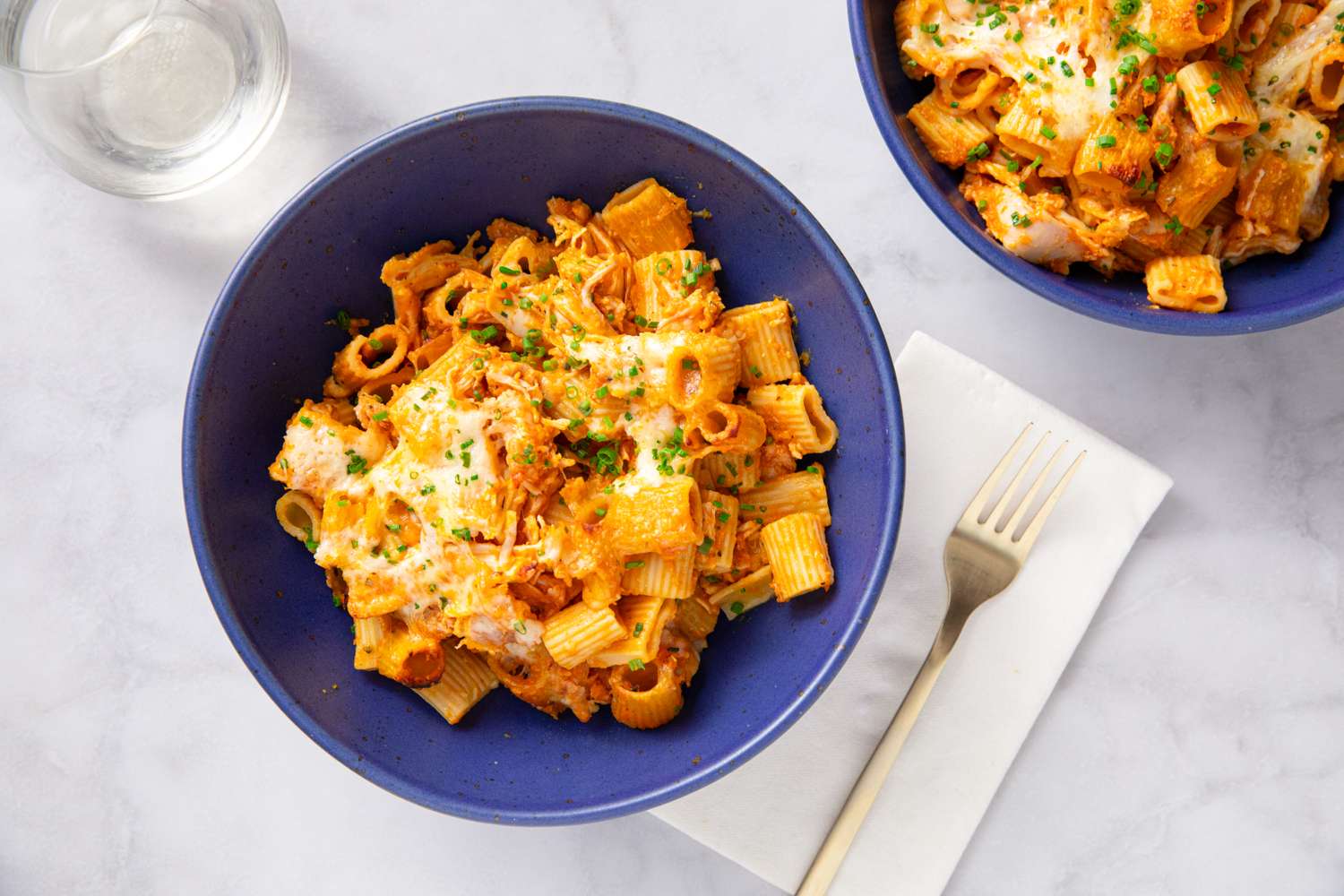 Pumpkin chicken pasta bake served in blue bowls placed on a white table with cutlery and a glass of water nearby
