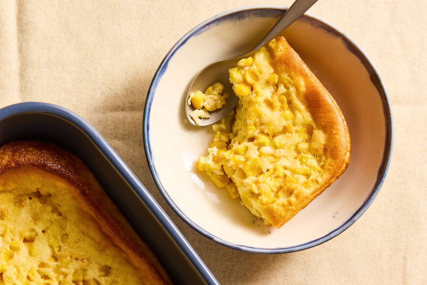 A portion of cornbread casserole served in a bowl with a spoon the main dish visible in a baking dish nearby