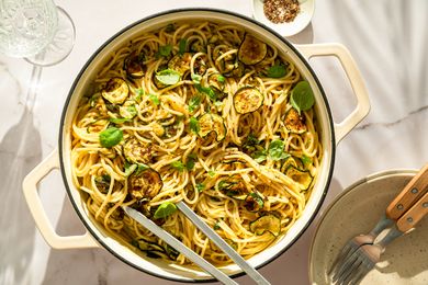A pot containing spaghetti with zucchini slices and garnished with basil, alongside a plate, utensils, and condiments