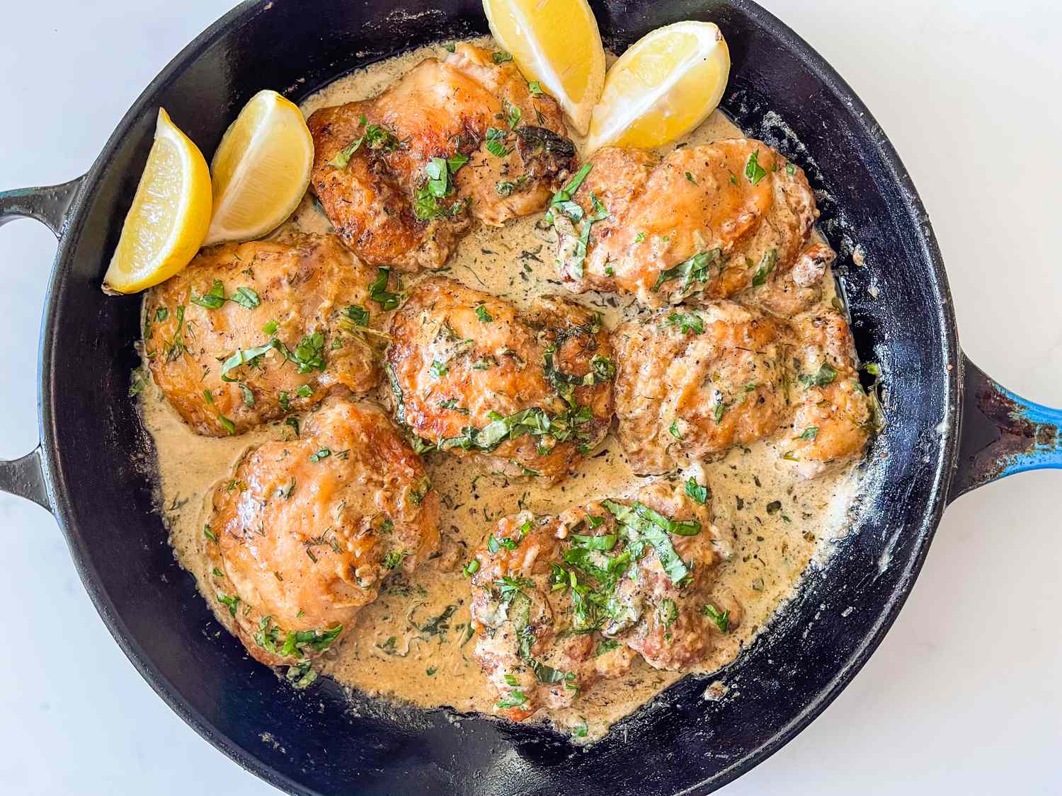 Cooked chicken thighs in a creamy herb sauce with lemon wedges in a cast iron skillet