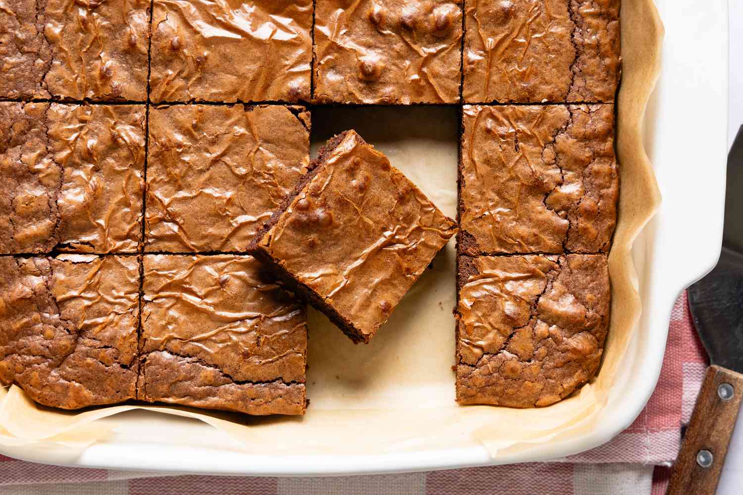 Overhead view of a white baking dish of brownies on parchment paper, sliced into squares with one slice removed