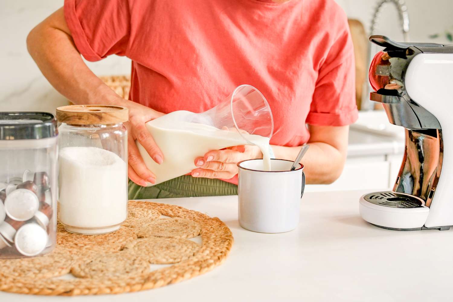 Person pouring milk into a mug near a countertop with coffee capsules sugar jar and an espresso machine