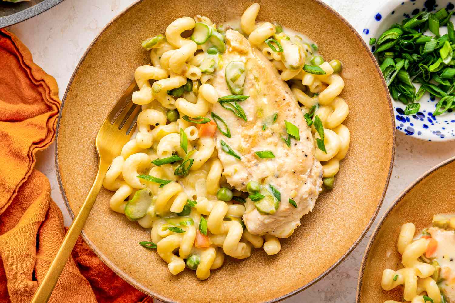 bowl of easy chicken primavera at a table setting with another bowl, a bowl of sliced scallions, and a table napkin