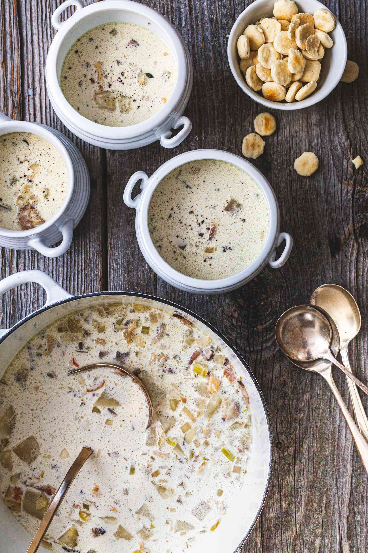 Classic New England Chowder in a Dutch Oven and in Individual Soup Bowls with Handles, Next to a Bowl of Oyster Crackers (with a Few Spilling onto the Table) and a Couple of Spoons