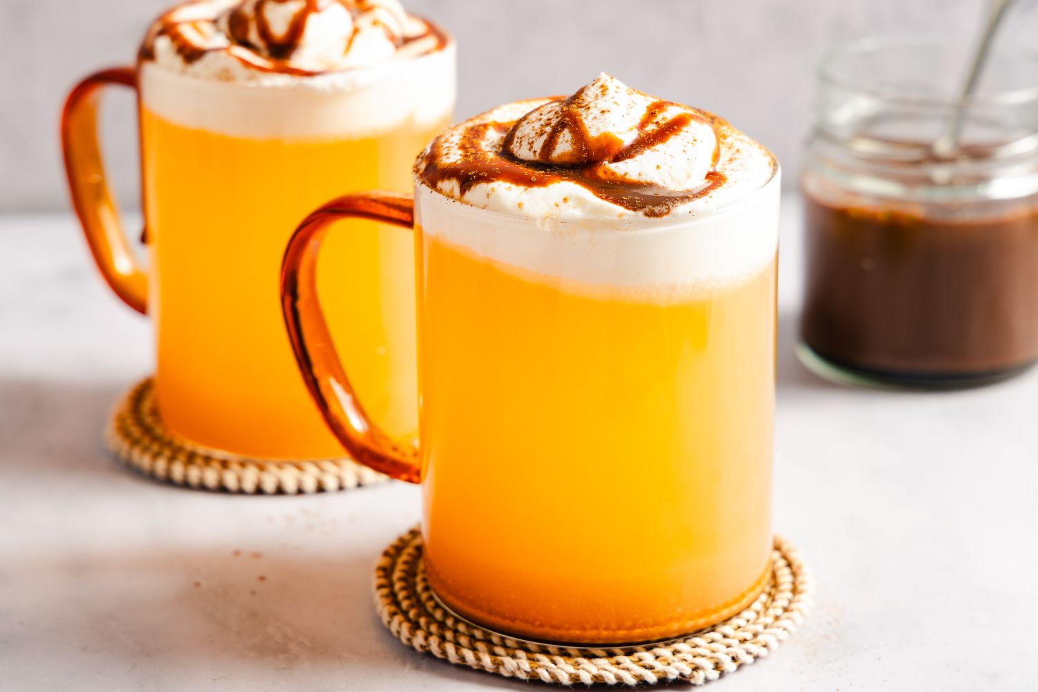Two mugs of caramel apple cider topped with whipped cream and caramel drizzle a jar of sauce in the background