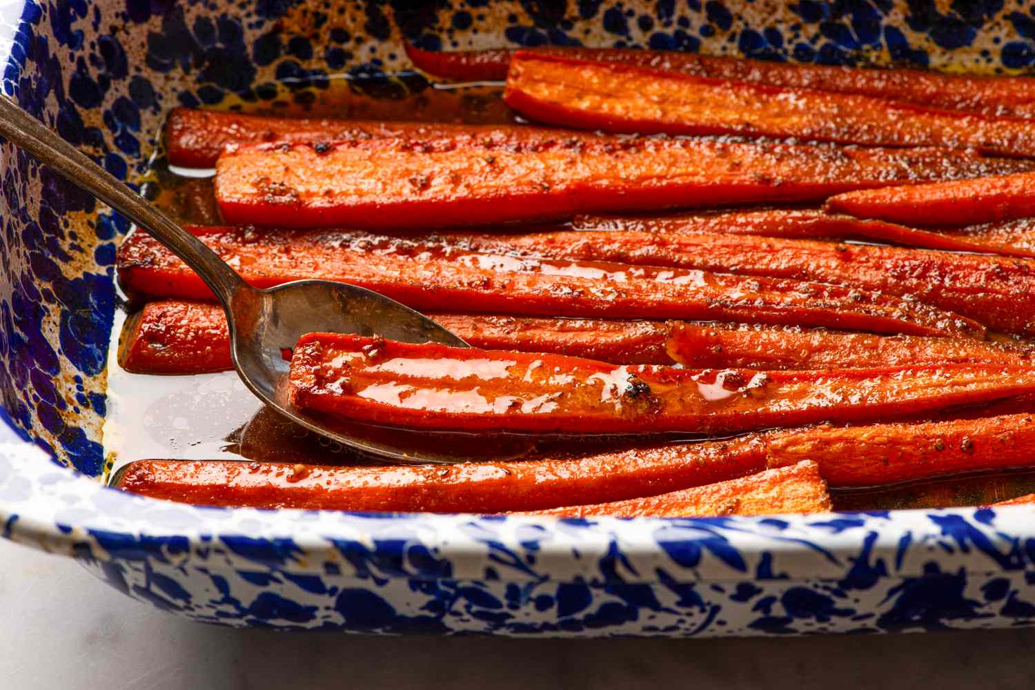 Cooked glazed carrots in a speckled blue baking dish with a spoon
