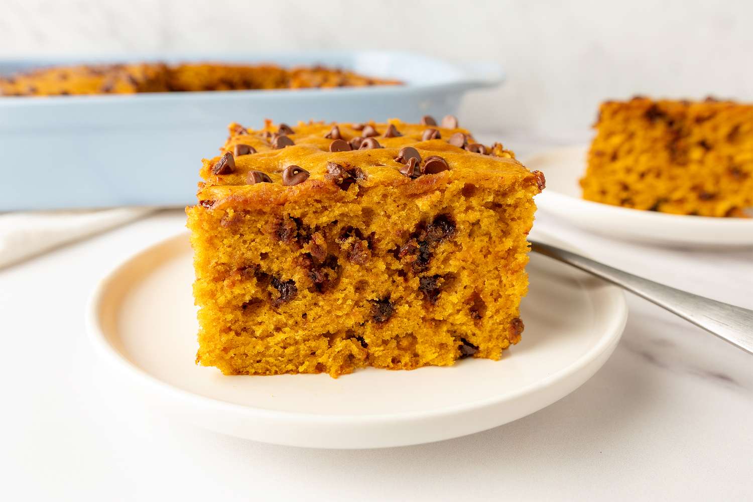 A slice of pumpkin chocolate chip cake on a white plate with a baking dish in the background