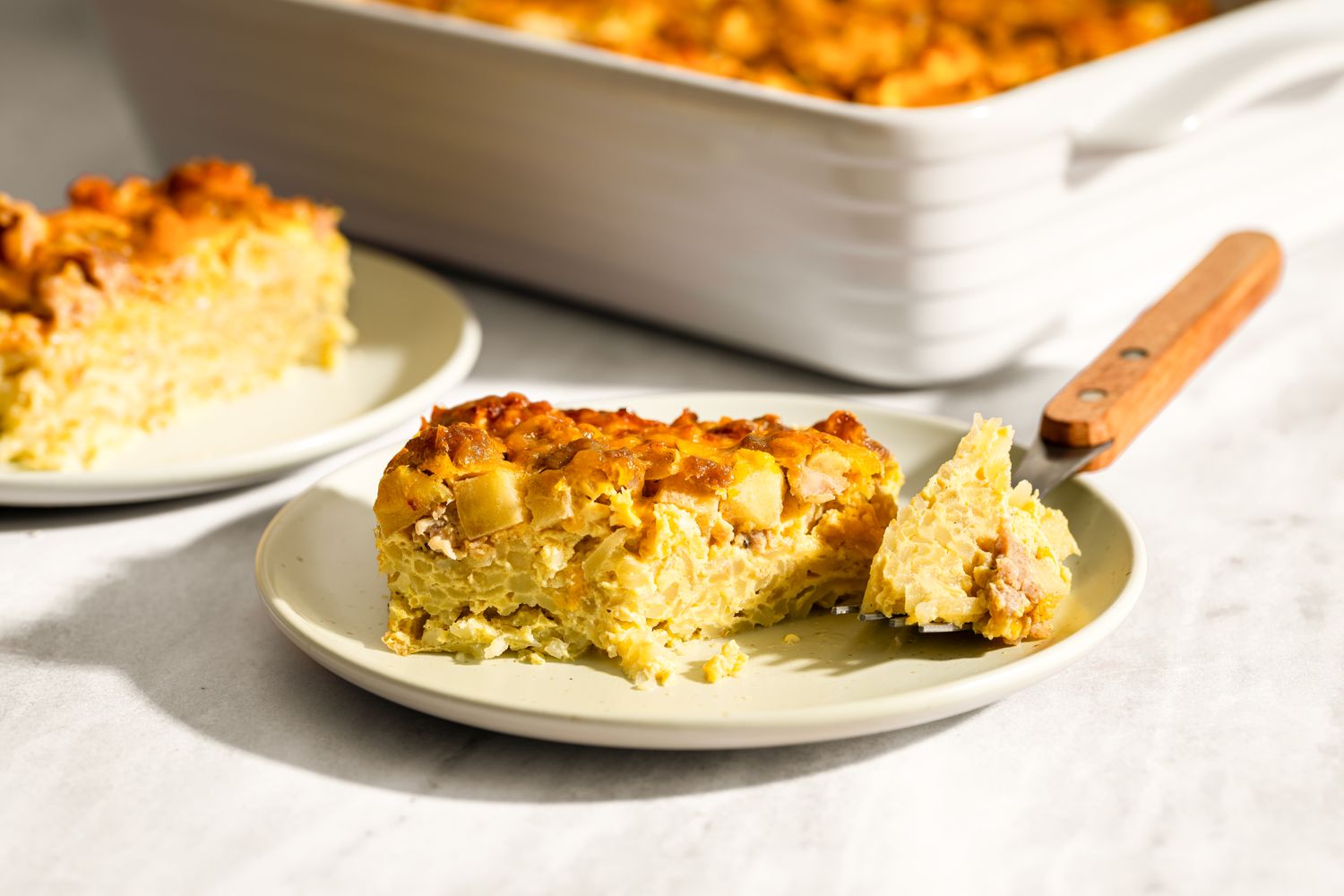 A plate with a slice of cheesy apple sausage casserole a casserole dish in the background