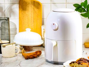 An air fryer on a kitchen counter surrounded by other kitchen tools like a set of olive wood measuring spoons, a cutting board, and two coffee mugs