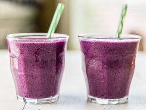 Two glasses of purple smoothies with green straws placed on a white surface