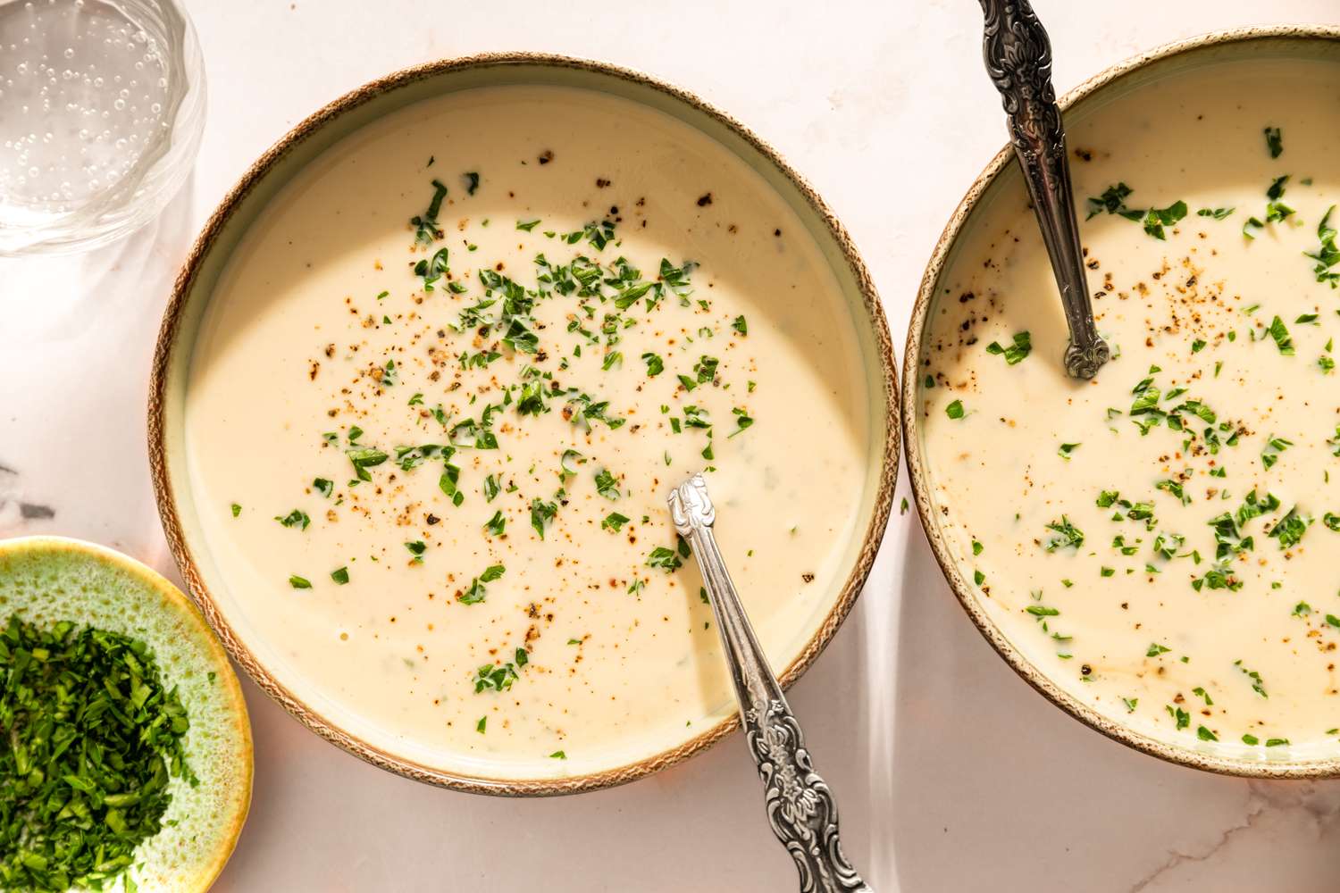Two bowls of cauliflower bisque garnished with herbs accompanied by a dish of chopped parsley and a glass of water