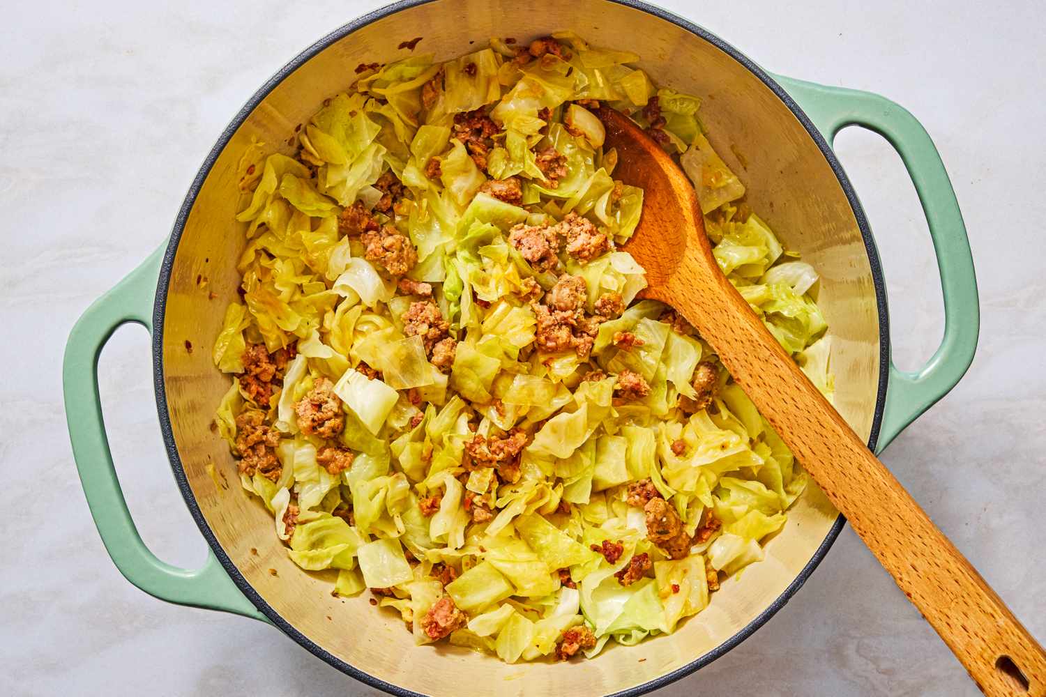 A pot containing cabbage wilting and some ground meat and a wooden spoon for the5-Ingredient Braised Cabbage “Lasagna” recipe part of a dish preparation