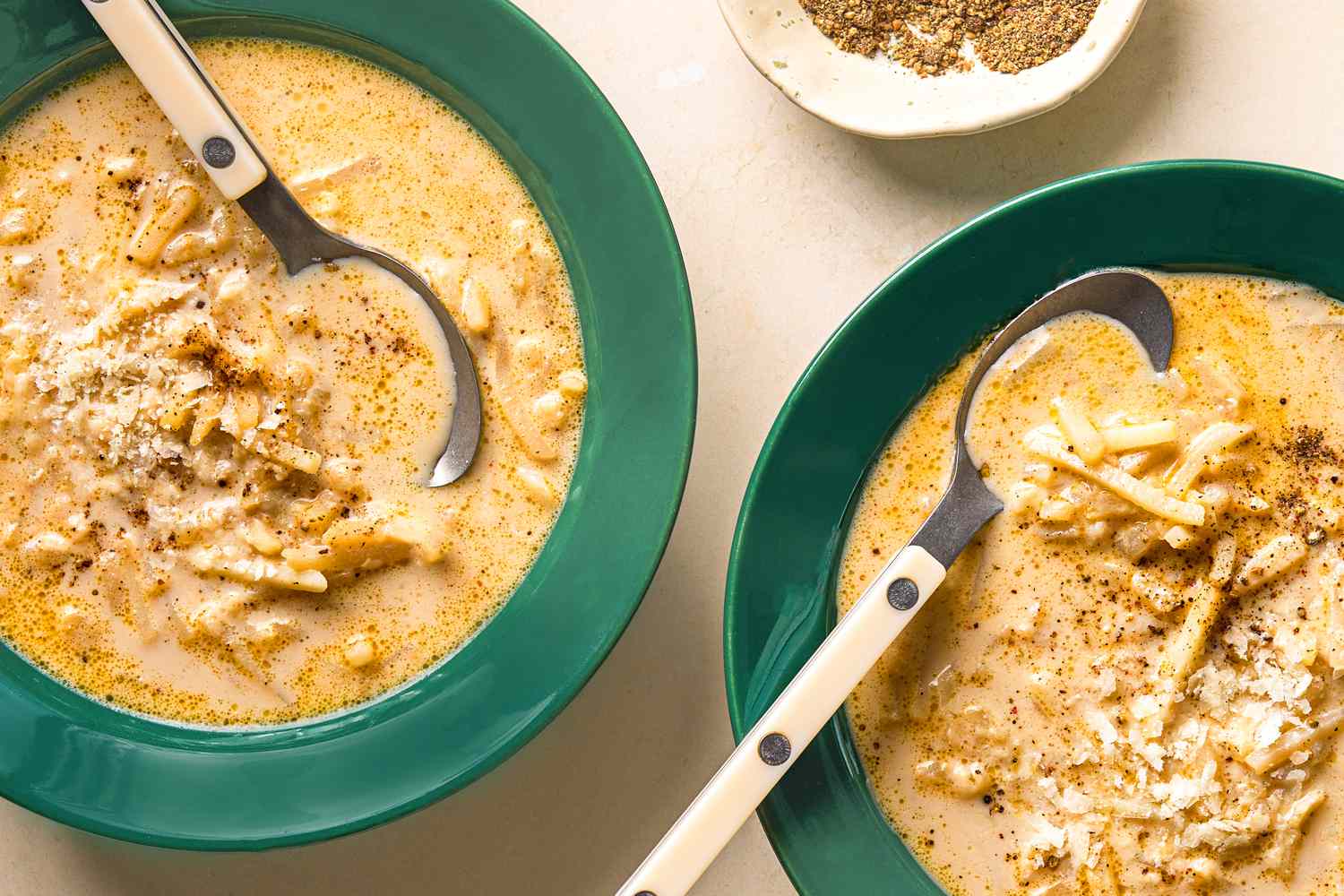 Two bowls of cacio e pepe potato soup with spoons topped with grated cheese and black pepper next to a small dish of black pepper