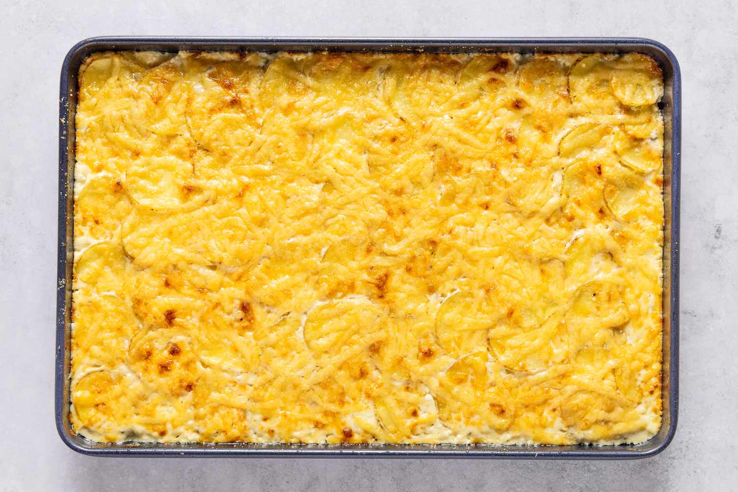 Overhead view of a 9x13 sheet pan of cooked potato and cheese mixture before garnishing with chives for Sheet Pan Au Gratin Potatoes recipe