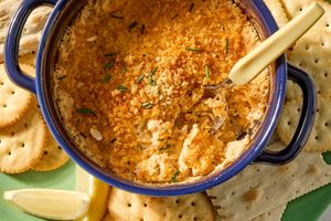 A casserole dish of baked lobster dip with bread crumb topping beside crackers and a lemon wedge