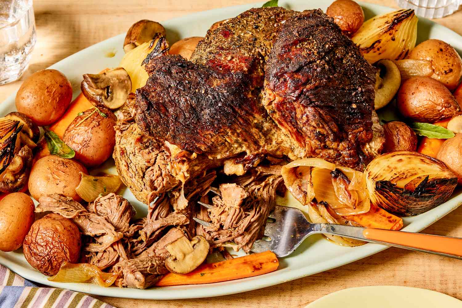 A slowcooked pot roast on a platter with vegetables like carrots potatoes and onions partially shredded with a serving fork